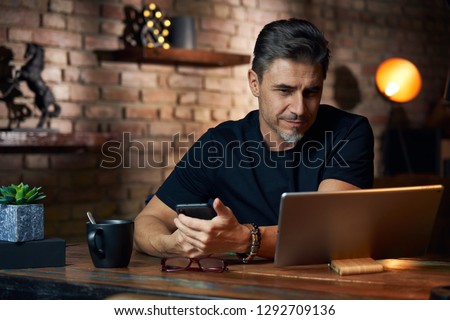 Older white man sitting at desk in dark room at home, working, using phone and tablet