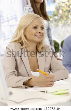 Portrait of attractive young businesswoman sitting at desk, writing notes, smiling.
