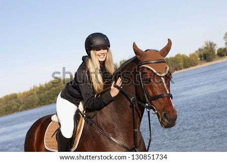 Happy female rider leaning over horse caressing back at riverside.