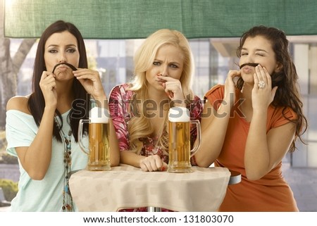 Attractive female friends having fun, forming moustache from hair, drinking beer.