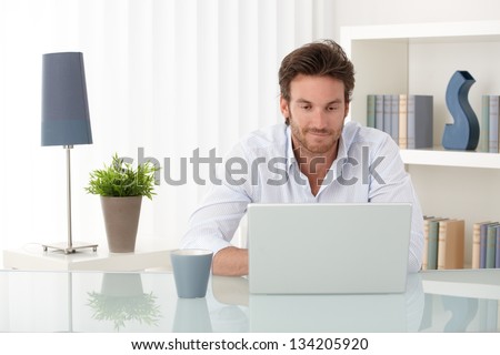 Handsome sitting at living room table, using laptop computer at home, smiling, looking at screen.