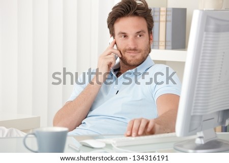 Portrait of smiling man speaking on mobile phone, sitting at desk, looking at computer screen.
