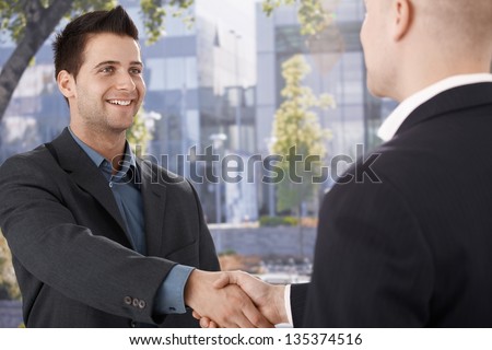 Businessmen shaking hands in front of office building, smiling.
