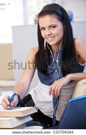 Pretty schoolgirl learning at home, smiling, looking at camera, writing notes, using laptop.