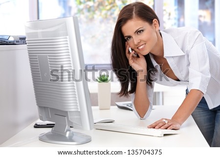 Beautiful young woman working in bright office, using mobile, smiling.