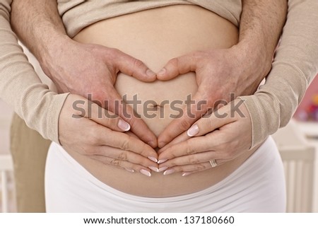 Closeup photo of naked pregnant tummy. Man\'s hands forming a heart around belly button.