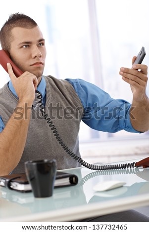 Determined businessman on landline phone call in office, with mobile phone handheld.