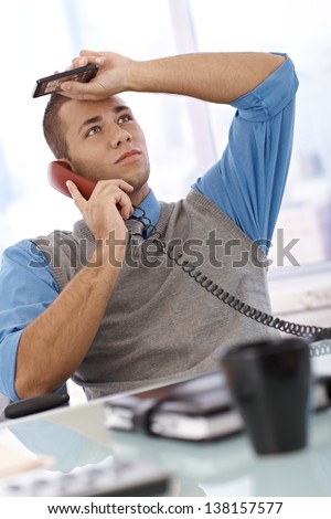 Troubled businessman at desk concentrating on landline phone call, holding mobile phone, looking up.