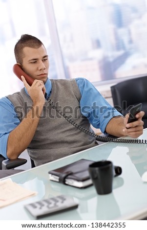 Businessman sitting in office busy using mobile phone texting, multitasking, on landline phone call.