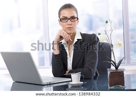Businesswoman at work, sitting in office with laptop computer, having coffee, holding pen, looking at camera.