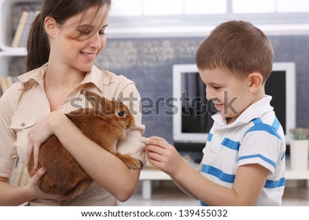 Smiling boy feeding cute pet rabbit handheld by happy young mother.