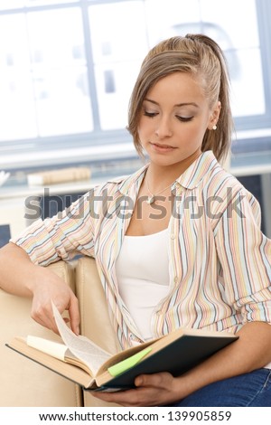 Blonde girl sitting at home, studying, reading a book.