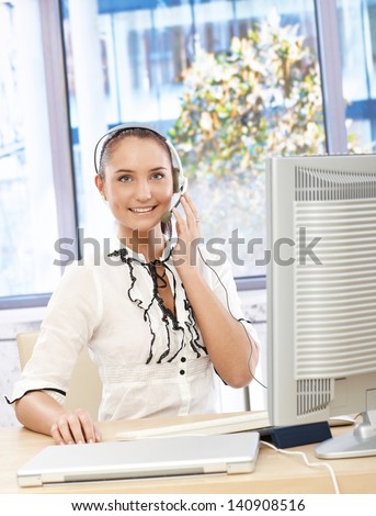 Happy office girl speaking on headset, smiling, using desktop computer in customer service office.