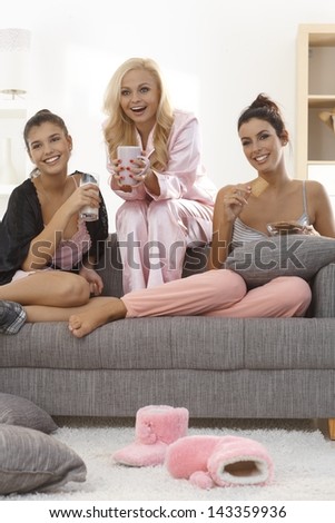Roommates sitting on sofa, wearing pyjamas, watching romantic movie on tv, smiling.