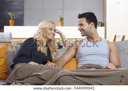 Young loving couple sitting on sofa at home, covered by blanket, smiling at each other with love.