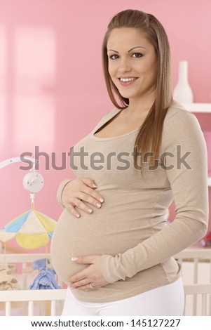 Portrait of happy pregnant woman standing in baby's room by crib, caressing belly, looking at camera.