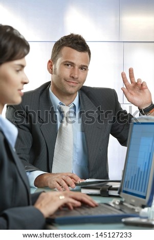 Young businessman raising hand at business meeting, sitting at table, looking at camera.