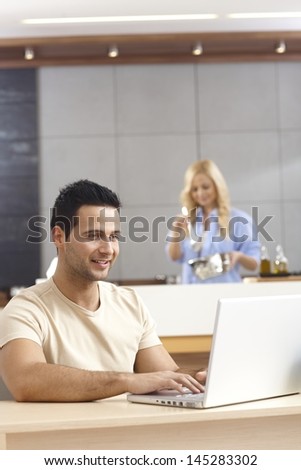 Young man sitting at table, using laptop computer at home, female cooking at background.