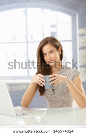 Portrait of businesswoman having coffee, sitting at table with laptop computer, smiling at camera.
