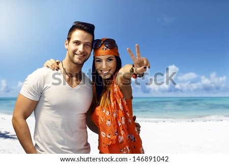 Happy Young Couple Embracing On The Beach, Smiling, Showing Victory Sign.
