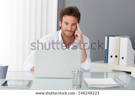 Businessman working at office, sitting at desk, looking at laptop computer screen, smiling, thinking.