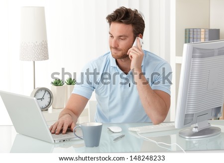 Casual businessman working at office desk, using mobile phone and laptop computer, typing, making phone call, smiling.