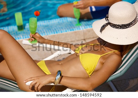 Sexy woman in bikini relaxing by pool, drinking cocktail, face covered with straw hat.