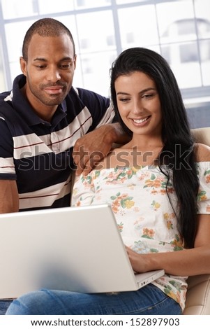 Attractive young mixed race couple browsing internet at home on laptop computer, smiling.