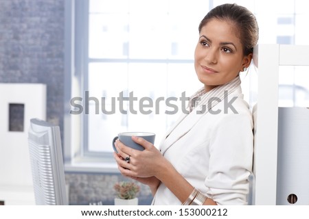 Portrait of attractive young businesswoman drinking tea in office, thinking, smiling, looking away.