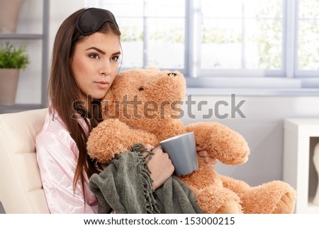 Morning portrait of sleepy young woman cuddling with teddy bear and blanket, holding coffee mug, wearing eye cover and pyjama, sitting in living room.
