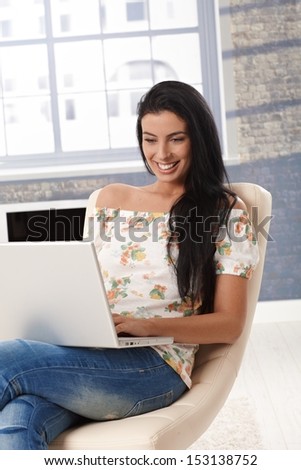 Happy young woman sitting at home, browsing internet on laptop computer, smiling.