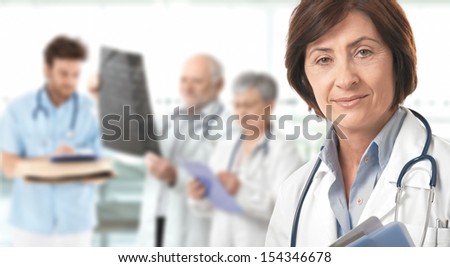 Portrait of senior female doctor looking at camera, medical team working in background.