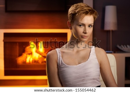 Attractive young woman sitting by fireplace at home, looking at camera.