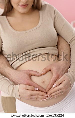 Closeup photo of naked pregnant tummy. Young couple forming a heart around belly button.