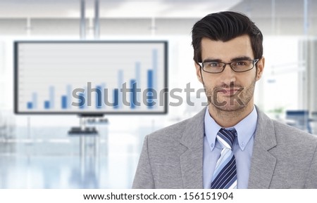 Portrait of confident young businessman at office with financial graph on TV.