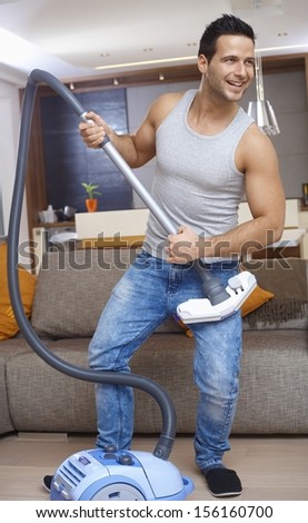 Young man holding vacuum cleaner as guitar, having fun at home, smiling.