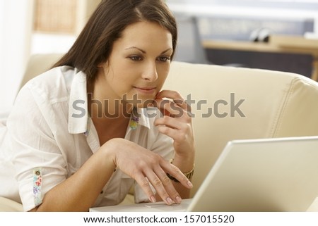 Young woman lying on sofa in living room, using laptop computer, browsing internet.
