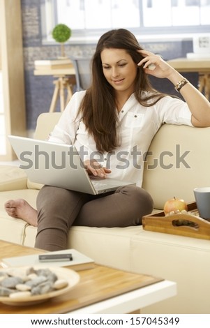 Young woman browsing internet on laptop computer at home on sofa, smiling.