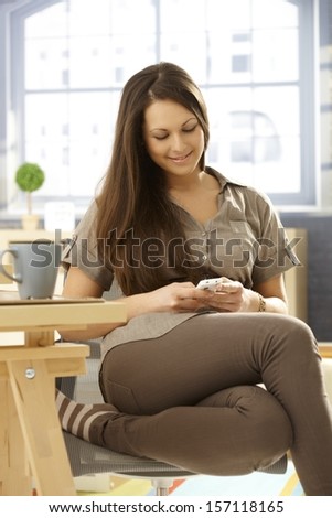 Young woman using mobilephone at home, sitting on chair, writing text message, smiling happy.