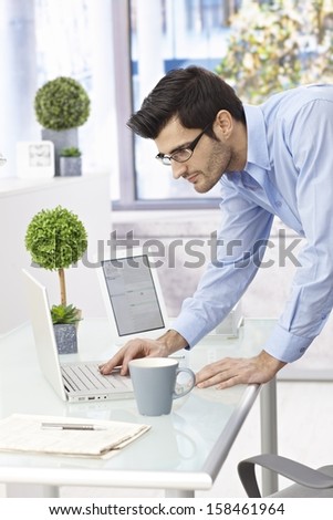 Young businessman using laptop computer standing at desk, leaning on it.