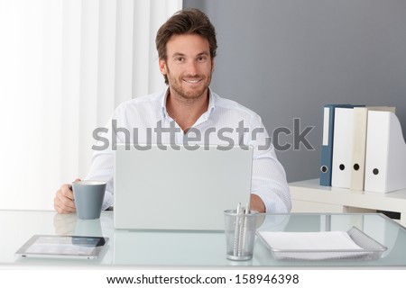 Happy Businessman Sitting In Office, Working With Laptop Computer, Having Tea, Smiling At Camera.