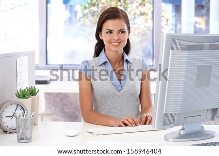 Smiling female casual office worker sitting at desk in bright office, using computer.