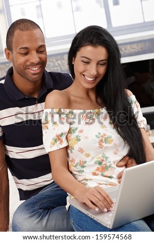Happy mixed couple using laptop computer at home, smiling, browsing internet.