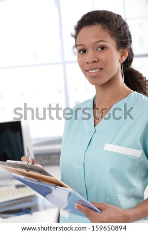 Young ethnic nurse holding case sheets, smiling.