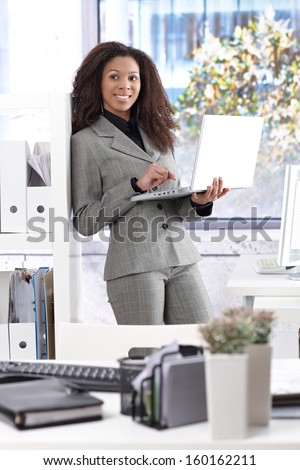 Pretty afro-american businesswoman leaning to bookshelf. working with laptop computer, smiling.