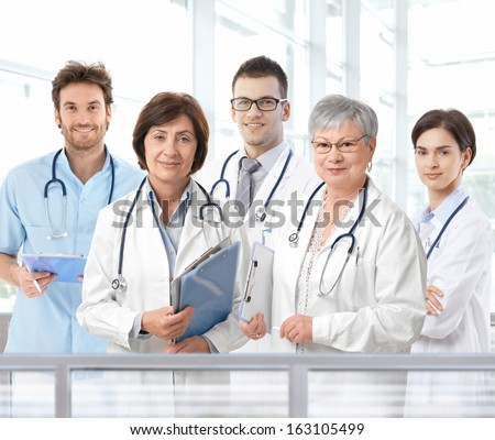 Team portrait of mixed aged medical doctors standing in hospital lobby, looking at camera, smiling.