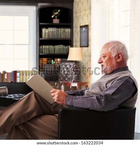 Elderly man sitting in armchair reading book at study at home.
