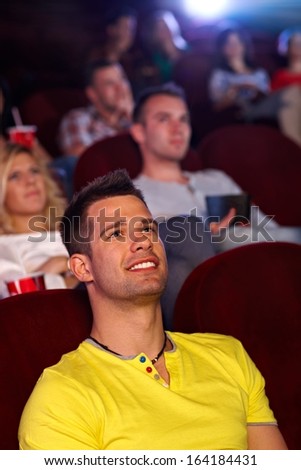 Young man sitting in multiplex movie theater, watching movie, smiling.
