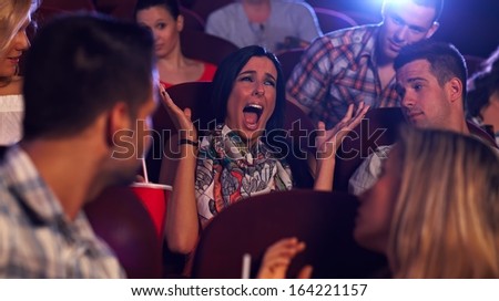Young woman screaming with horror in movie theater, others watching.