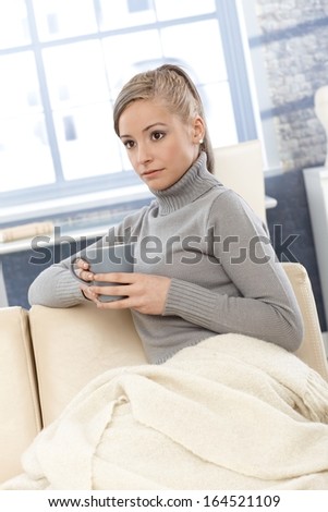 Young woman drinking tea at home, covered by blanket.